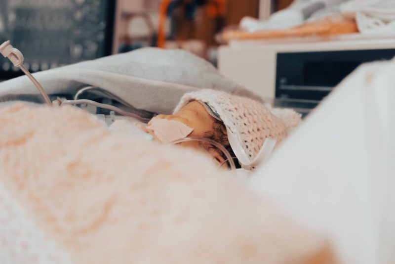 medical mom |  Epidermolysis bullosa News |  Baby Izzy sleeps in the NICU while attached to multiple tubes and machines.  She wears a small pink knit hat and is covered with a matching blanket.