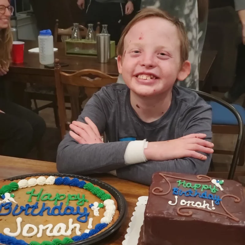 miracle | Epidermolysis Bullosa News Today | Jonah smiles for the camera while sitting at the table behind two large birthday cakes on his 13th birthday last Sunday