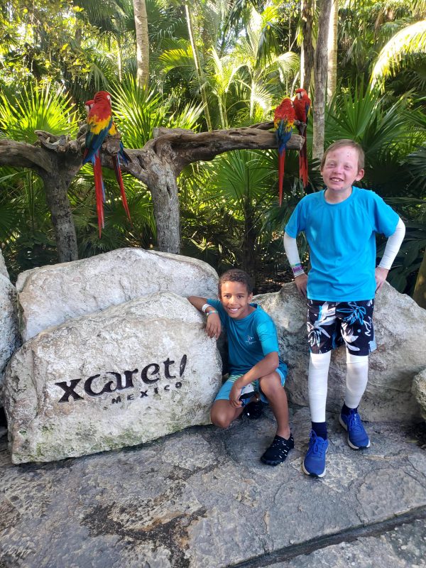 good times with EB | Epidermolysis Bullosa News | Gideon and Jonah pose next to a large rock that's engraved to read "Xcaret Mexico!" Behind the boys are palm trees and several macaws perched on a tree branch.