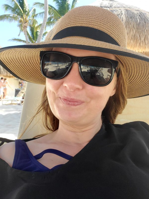 good times with EB | Epidermolysis Bullosa News | Patrice takes a selfie while relaxing on a lounge chair on the beach in Mexico. She's wearing dark sunglasses and a floppy hat.