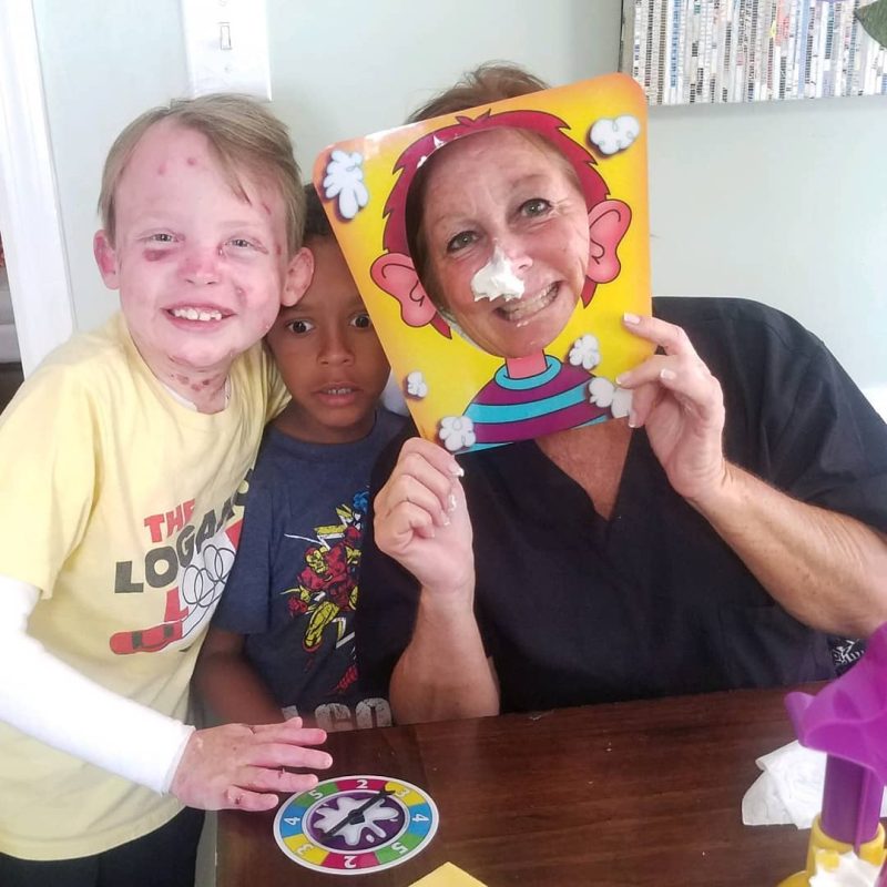 caregiver nurse | Epidermolysis Bullosa News | from left, Jonah, Gideon, and Clair indoors at a table. Clair is holding a Pie Face mask to her face and has cream on her nose