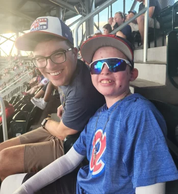 moments of joy | Epidermolysis Bullosa News | Jonah and his dad, Matt, smile from their seats at an Atlanta Braves baseball game. Jonah is wearing a Braves T-shirt, sunglasses, and a baseball cap