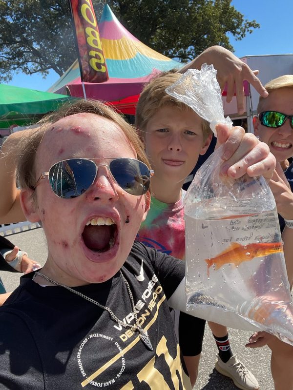 A selfie of Jonah shows him holding his newly won goldfish in a bag at the fair. Jonah is wearing dark sunglasses and a black T-shirt and is clearly screaming in victory. Behind him are his school friends. 