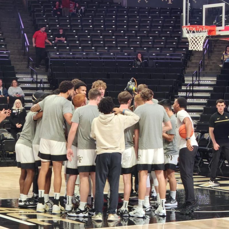 About 10 men wearing gray T-shirts and white shorts with black bands at the bottom huddle in the lane near a basket on a basketball court. A few other males are around the men. In the background are the stands, with about five people in them.