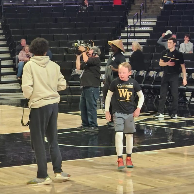 A boy walks off a basketball court stands just outside the lane on a basketball court. The boy is dressed in a black and gold Wake Forest University T-shirt, shorts, and orange sneakers. He is wearing white protective bandages or sleeves on his arms and legs. A man with a cameraman is behind him, and another man in a gray sweatshirt and black pants is on the court. A few people are in the stands.