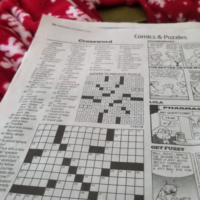 A close-up photo of a crossword puzzle next to comics in what appears to be a newspaper. A fuzzy red and white blanket is visible in the background.