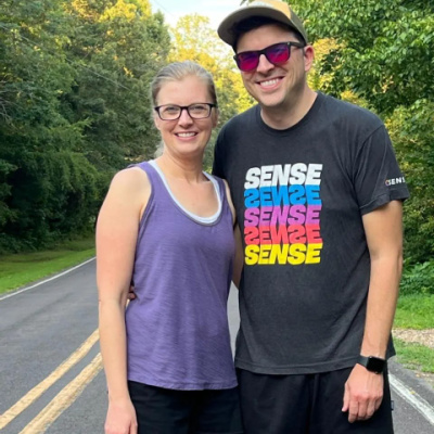 A woman and man smile and pose outside with one arm around each other. They're standing on a small, two-lane road surrounded on both sides by tall, thick, green trees. The woman is wearing a purple tank top and glasses, while the man is wearing a baseball cap, shaded glasses, and a gray graphic T-shirt.