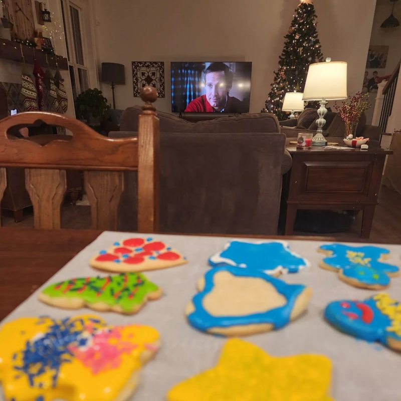 A dark photo of a living room decorated for Christmas, with a Christmas tree in the background and a television screen on. In the foreground are freshly baked and decorated Christmas sugar cookies with various colors of frosting and sprinkles.