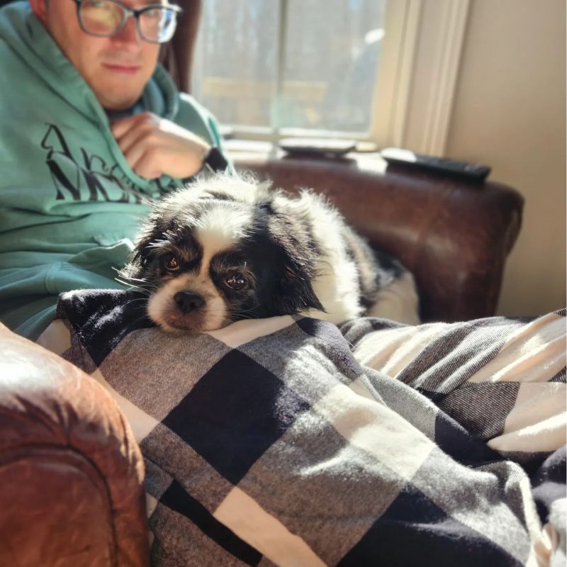 A man wearing large glasses and dressed in a green hoodie and pajama bottoms sits in a reclining chair with a dog on his lap.
