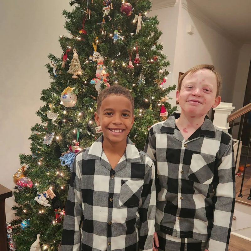 Two young brothers stand in front of a decorated Christmas tree, both wearing plaid pajamas.
