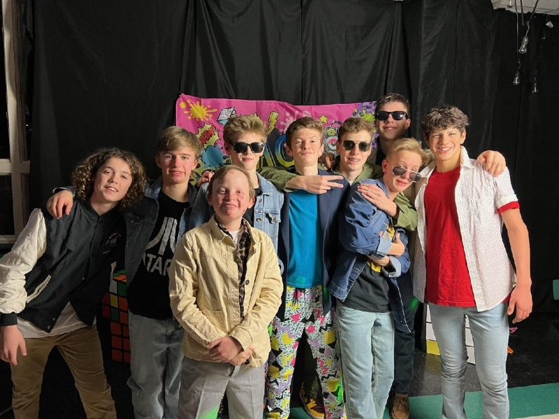 A group of nine eighth grade boys pose with their arms around each other at a school dance. A few boys are wearing aviators, and one is making a peace sign with his right hand. They're standing in front of a black curtain and a pink banner.