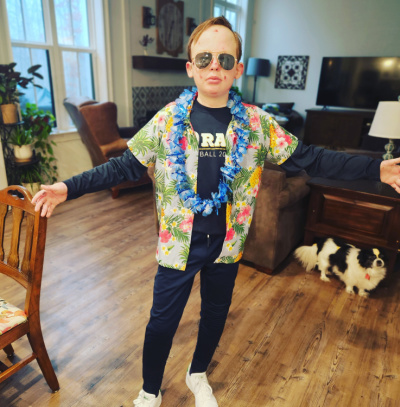 An eighth grade boy poses with his arms out in his living room. He's wearing aviators, a Hawaiian shirt, a blue lei, jeans, and white sneakers. A small black and white dog is visible just behind him.