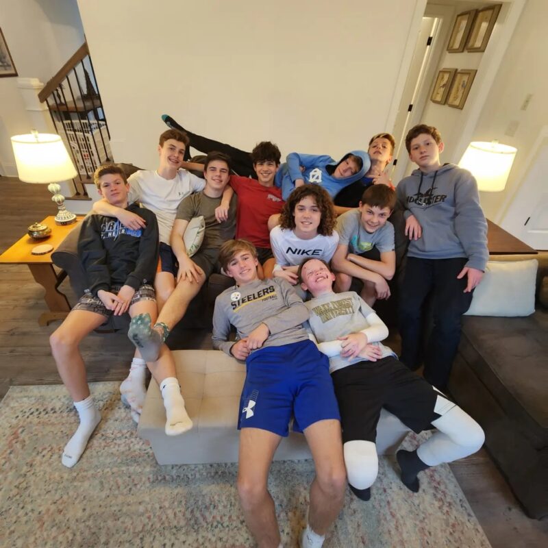 A group of 11 teenage boys in various sports clothing cram together on a living room couch for a group photo. 