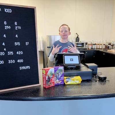 A teenage boy flashes some type of hand signal while standing behind a cash register at a coffee shop.