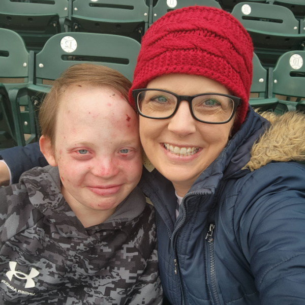 A mom and her teenage son smile for a selfie while seated in the stadium. The mom is wearing a red knit hat, glasses, and a puffy blue coat, while her son is wearing a camo sweatshirt. 