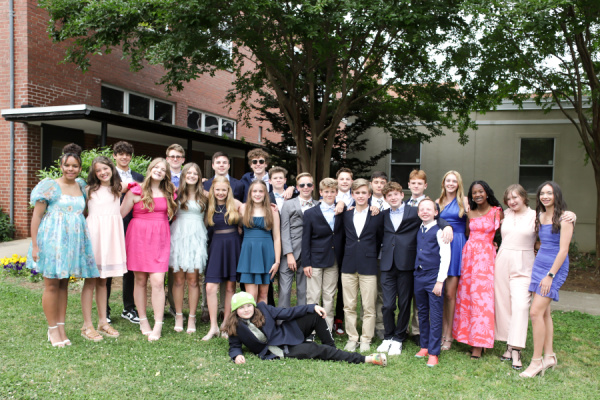 About 25 teenagers of various heights pose as a group in mostly formal attire on a lawn in front of what appears to be a school building. All are standing, aside from one girl lying on the ground at center. 