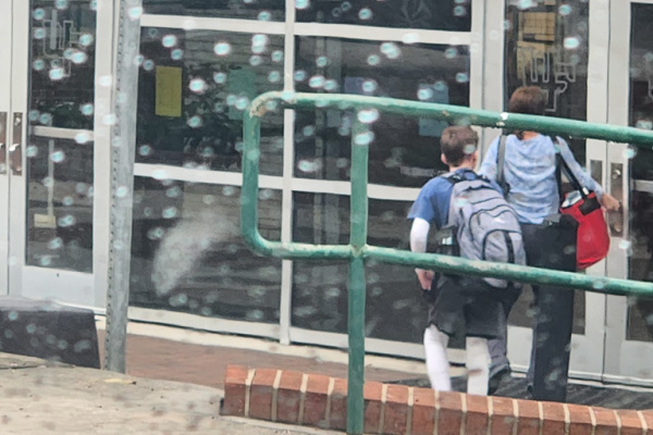 A photo taken through a car window with rain droplets on it shows a small teenage boy's back as he walks into a high school. The boy is wearing a blue T-shirt, has white bandages covering both arms, and has a large gray backpack. Another student in front of him is opening the door. 