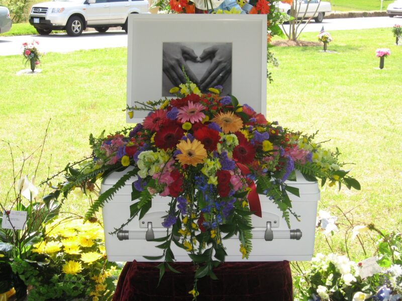 A tiny white casket sits in front of a new grave site in a cemetery. An arrangement of flowers is placed on top of the baby's casket.