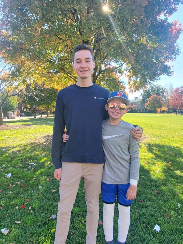 On a bright and sunny day, two boys, one much taller than the other, stand with their arms around each other and smile for the camera. They're standing in what looks like a big park, with several trees that are changing colors, indicating it may be the start of autumn. Rays of sunshine filter through the tree behind them. The shorter of the two, who has EB, is wearing a blue baseball cap and sunglasses, and his legs are entirely covered with protective bandages. 