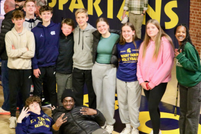 A group of teenagers pose for a photo at a school basketball game. Nine stand in a row, while two lie on the floor in front of them. A black banner in the background is only partially visible but appears to read "Spartans."
