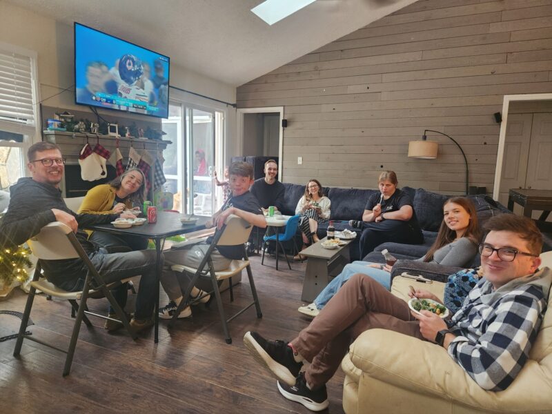 An extended family of eight sits around a folding table and on two couches in a large living room. They're eating a Thanksgiving meal. A Bears game is on the television screen above them. 