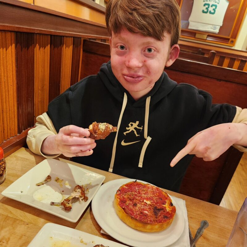 A teenage boy smiles while eating at a restaurant. He has two plates in front of him: one with a small deep-dish pizza, and the other with bones from chicken wings. He's holding the last wing in his right hand, and pointing at the pizza with his left. He has reddish hair and is wearing a dark hoodie.