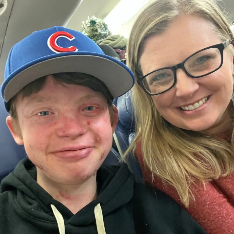 A blond woman and her teenage son smile for a selfie while seated next to each other on an airplane. The woman is wearing dark-framed glasses and a scarlet sweater, while her son is wearing a hoodie and a Chicago Cubs baseball cap.