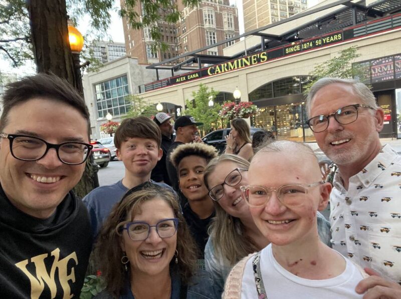 Two families squeeze into the frame on the sidewalk outside of Carmine's restaurant in Chicago. There are two sets of parents, two boys, and one girl. 