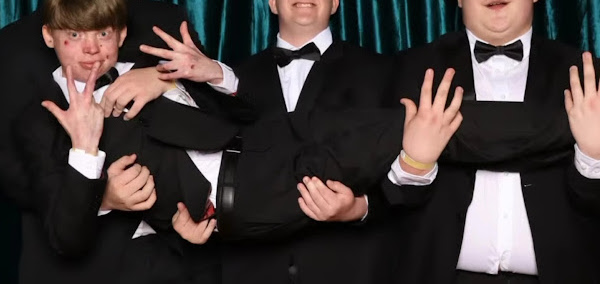 A horizontal color photo of three high school boys acting silly. They're all wearing black tuxes or suits, and we can only see the face of the boy on the left, who is gesturing with both hands. The faces of the other two boys are cut off, so we can't see them. 