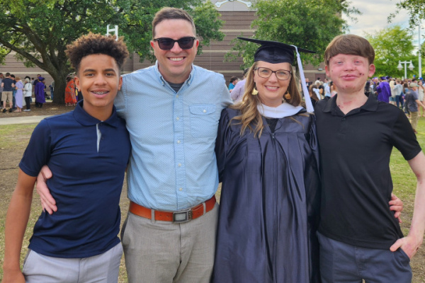 A family of four — mom, dad, and two teenage boys — smile for a photo while standing side by side. The mom is wearing a graduation cap and gown after earning her master's degree.