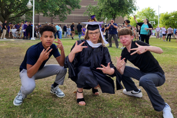 A mom poses for a silly photo with her two teenage sons outside on the grass. She's wearing a graduation cap and gown, and all three are squatting close to the ground and making signs with their hands.